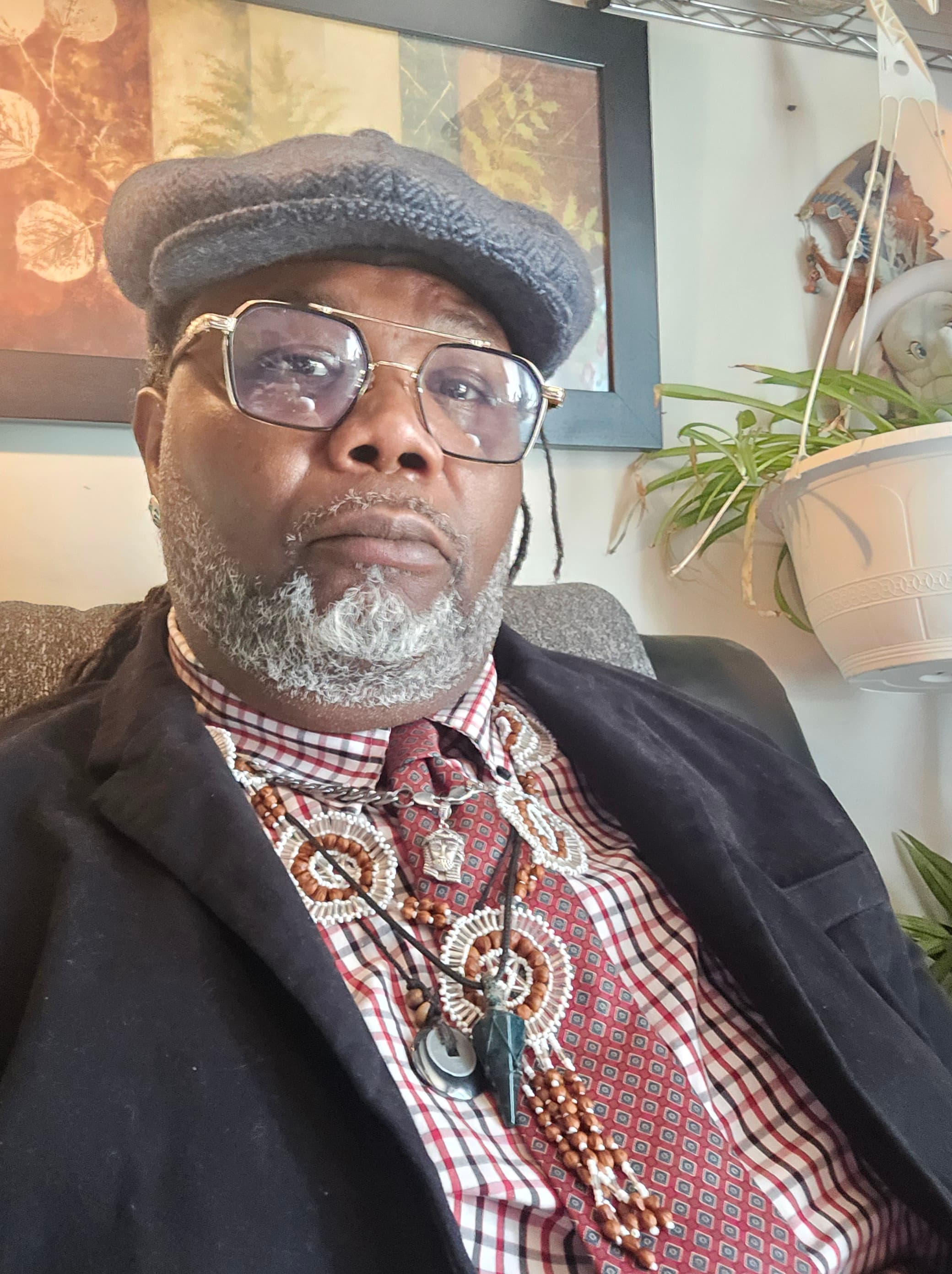 Man with a gray beard wearing a flat cap, glasses, and layered beaded necklaces.
