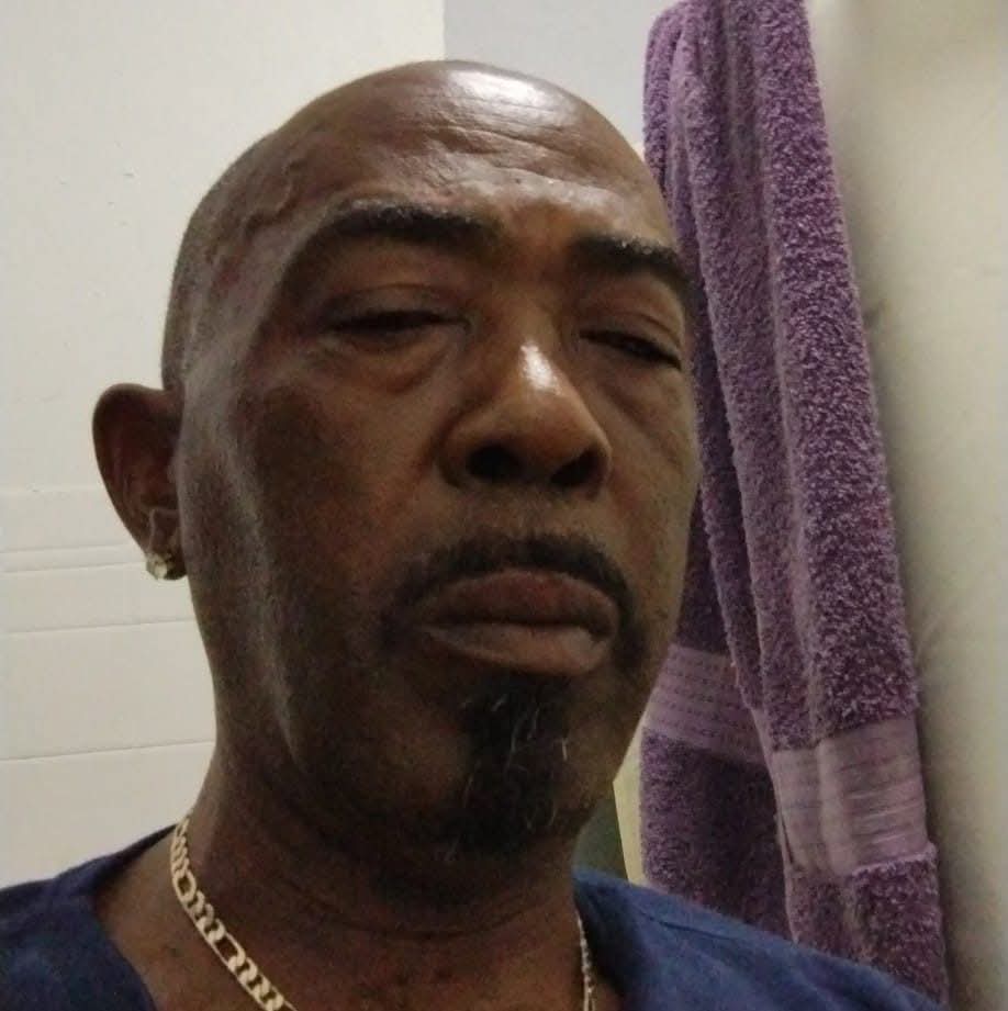 Bald Black man with a goatee and gold chain, posing near a purple towel.