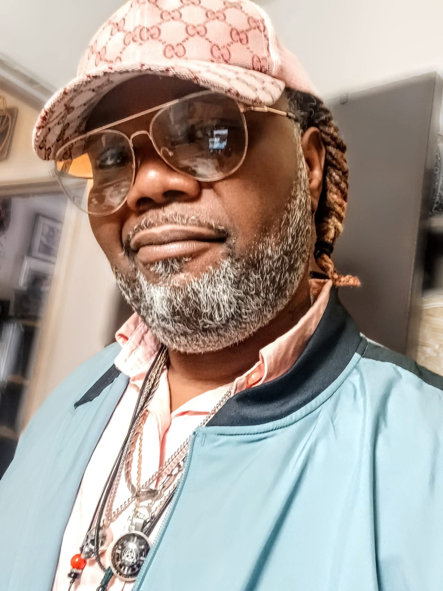 Man with salt-and-pepper beard, dreadlocks, aviator sunglasses, pink cap, and blue bomber jacket.