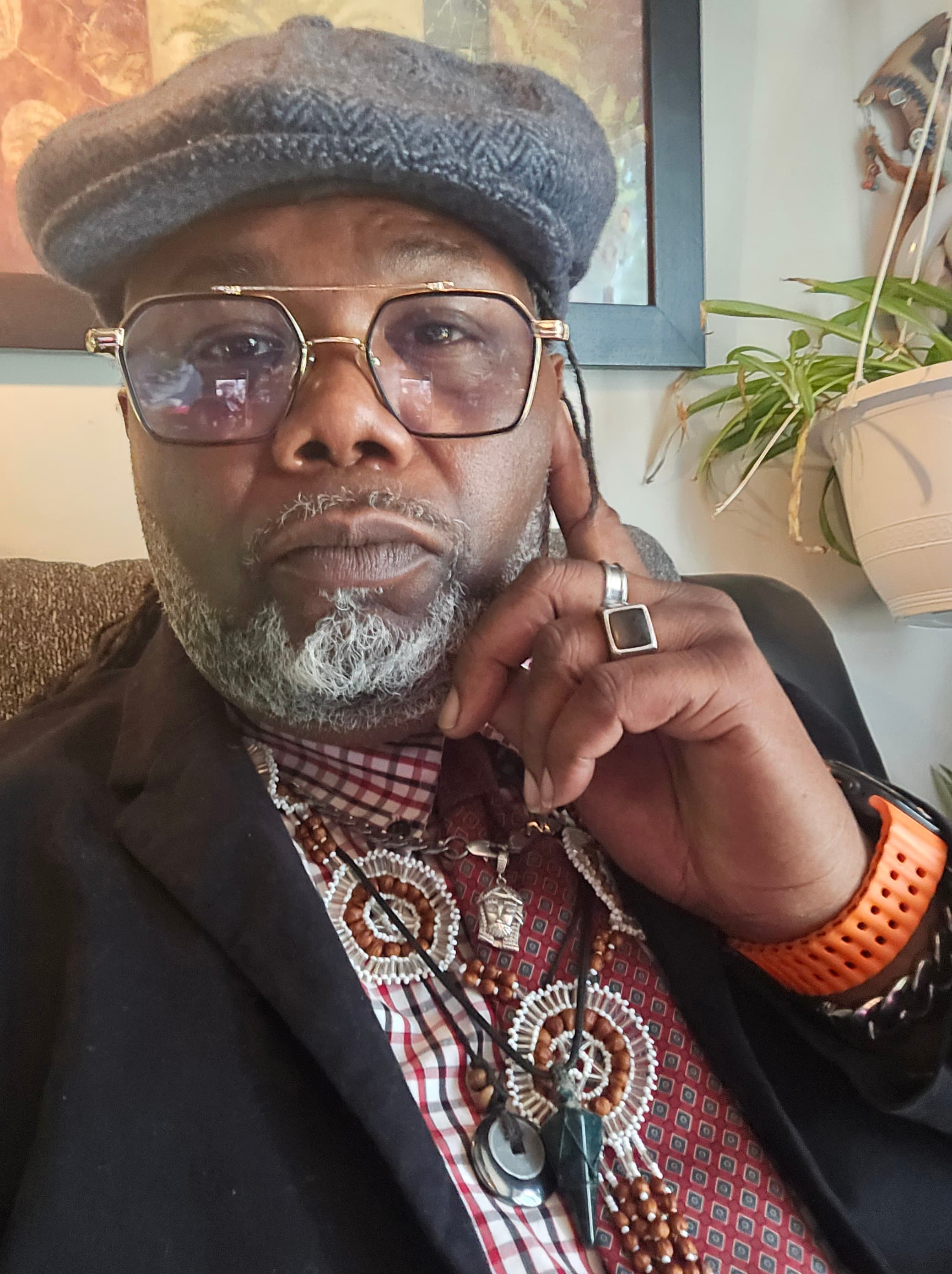 A Black man with a grey beard, flat cap, glasses, and several ornate necklaces.
